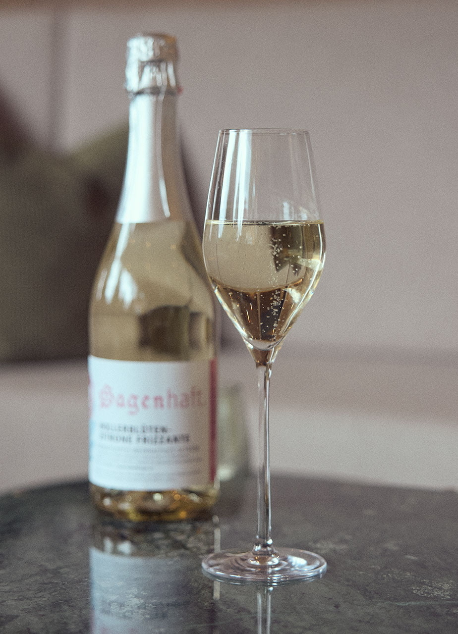 Ein Sektglas gefüllt mit Holunderfrizzante vor einer vollen Flasche desselben auf einem Beistelltisch stehend. Im Hintergrund ist eine Sitzbank in der Hotel Guglwald Lobby zu sehen.