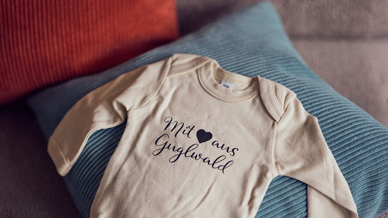 Ein Babybody mit der Inschrift mit Liebe aus Guglwald liegt auf einem Sofa